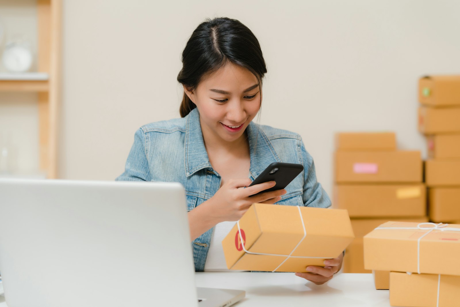 A small business owner checks a package with her phone beside a laptop and boxes.